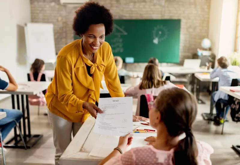 Happy African American teacher giving test results to elementary student in the classroom.